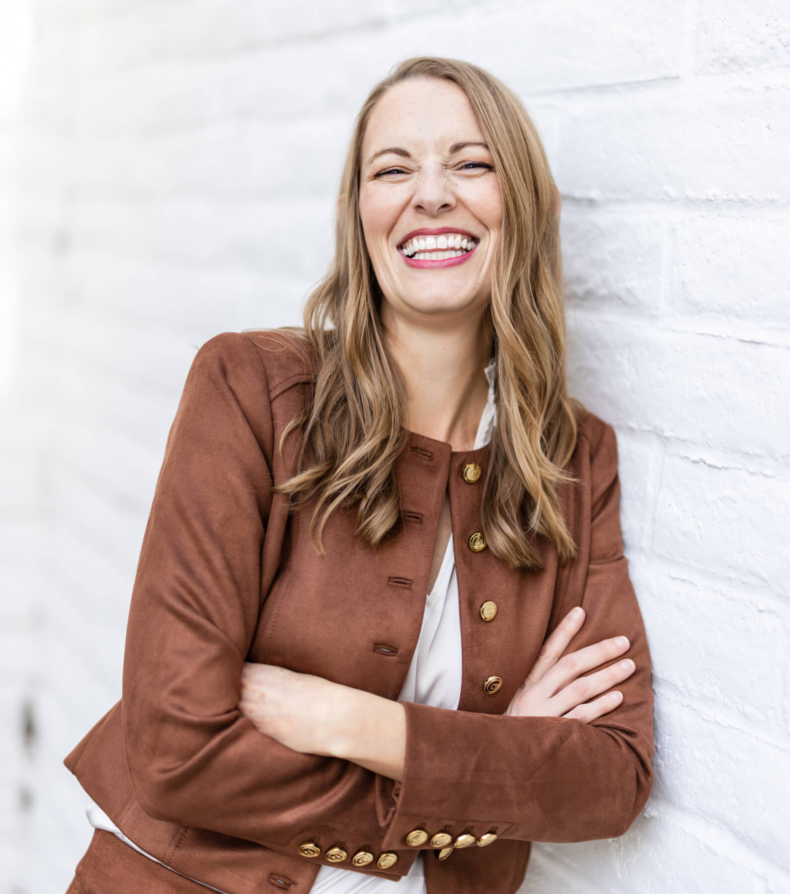 A woman with long, wavy hair, wearing a brown jacket, leans against a white brick wall with her arms crossed, smiling broadly.
