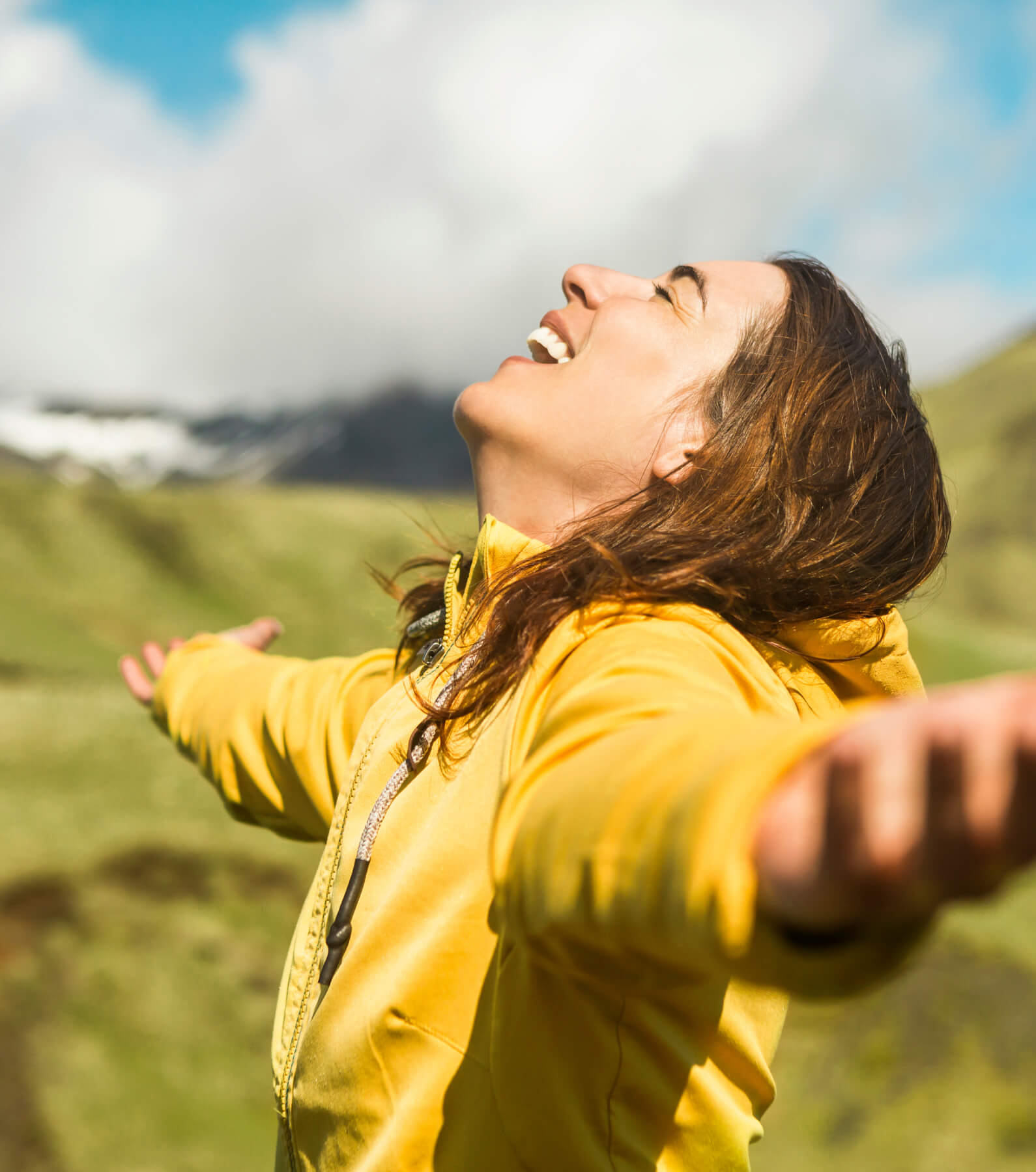 A person in a yellow jacket extends their arms wide and appears joyful in a green mountainous landscape under a partly cloudy sky.