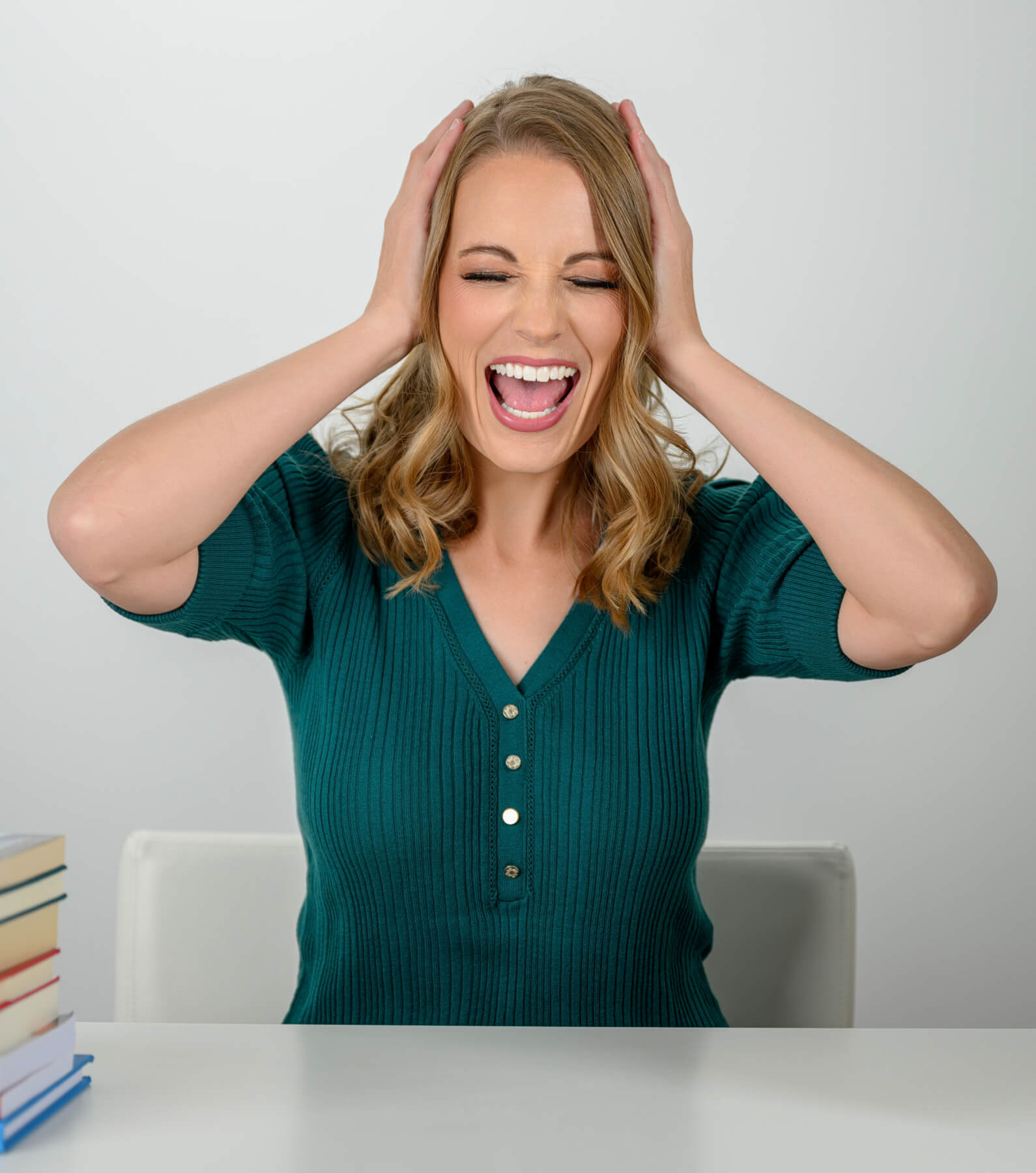 A woman with shoulder-length blonde hair, wearing a green sweater, sits at a table with a stack of books. She is holding her head with both hands and has her eyes closed while laughing.