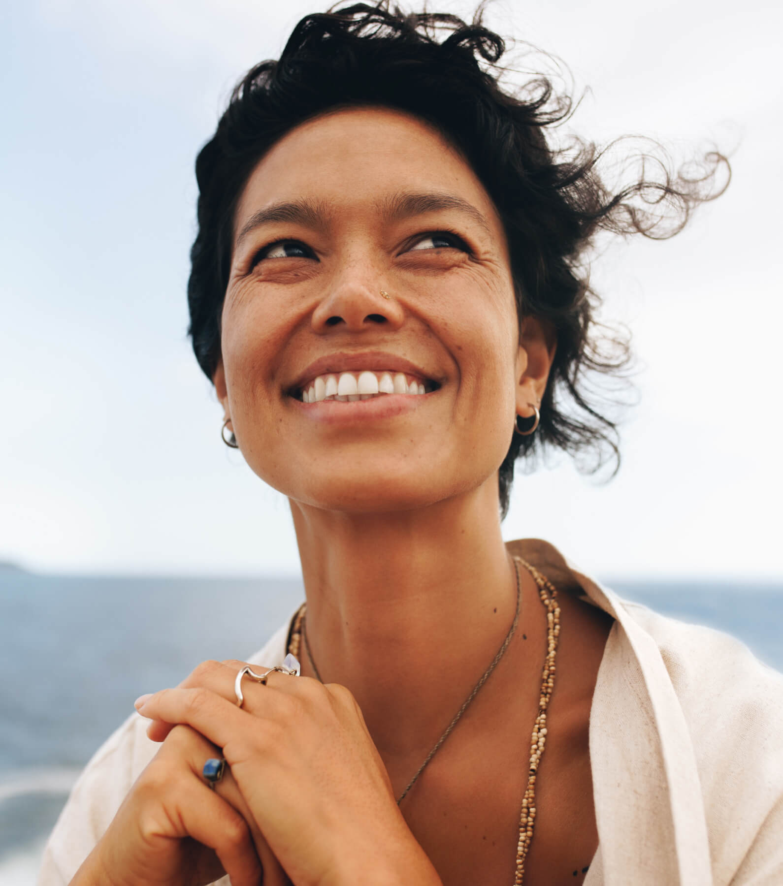 A person with short curly hair smiles while looking to the side, standing outdoors near the ocean.